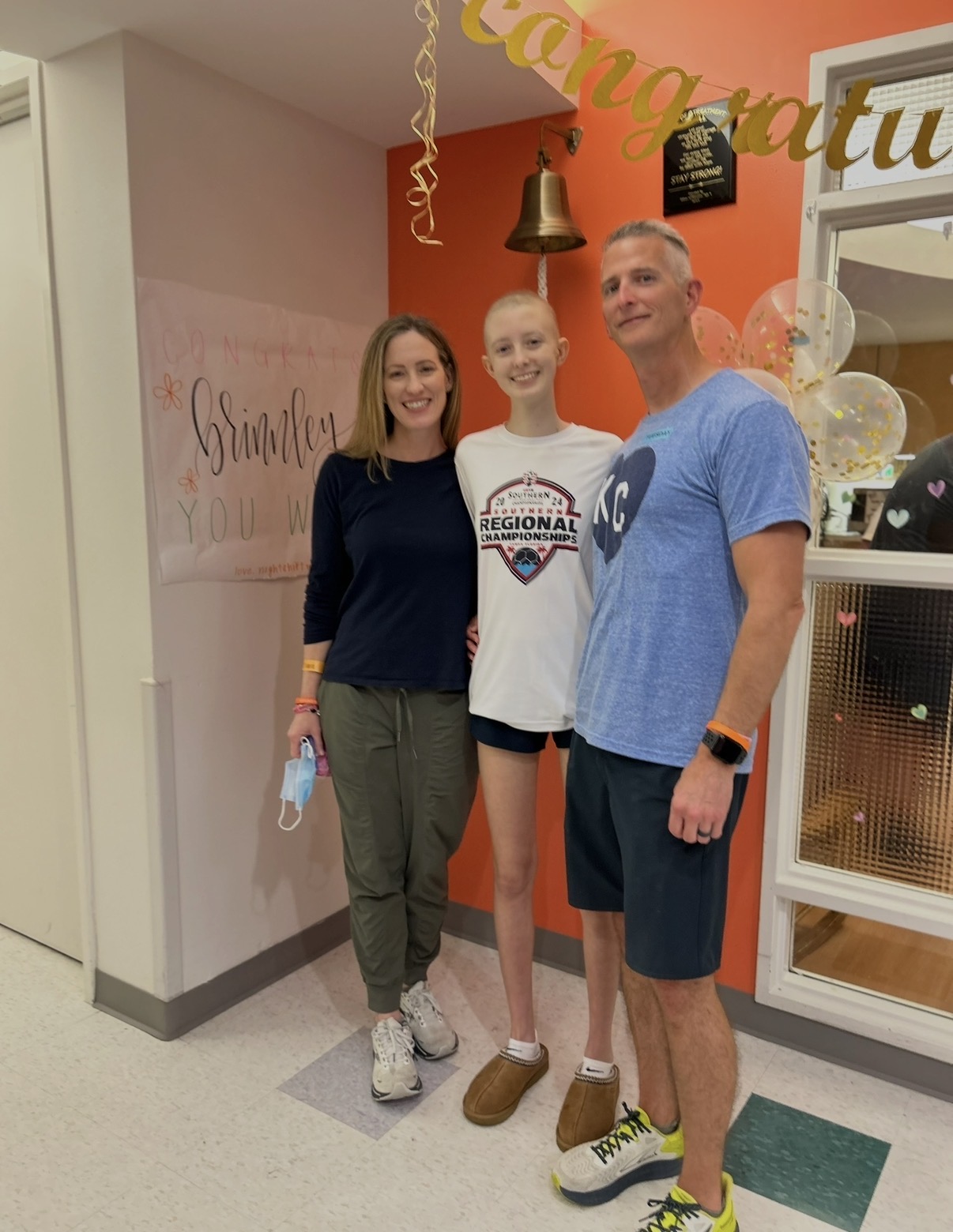 Brinn Lisenby smiling with her parents beneath the brass celebration bell in the hospital hallway after completing cancer treatment at Texas Children's Hospital