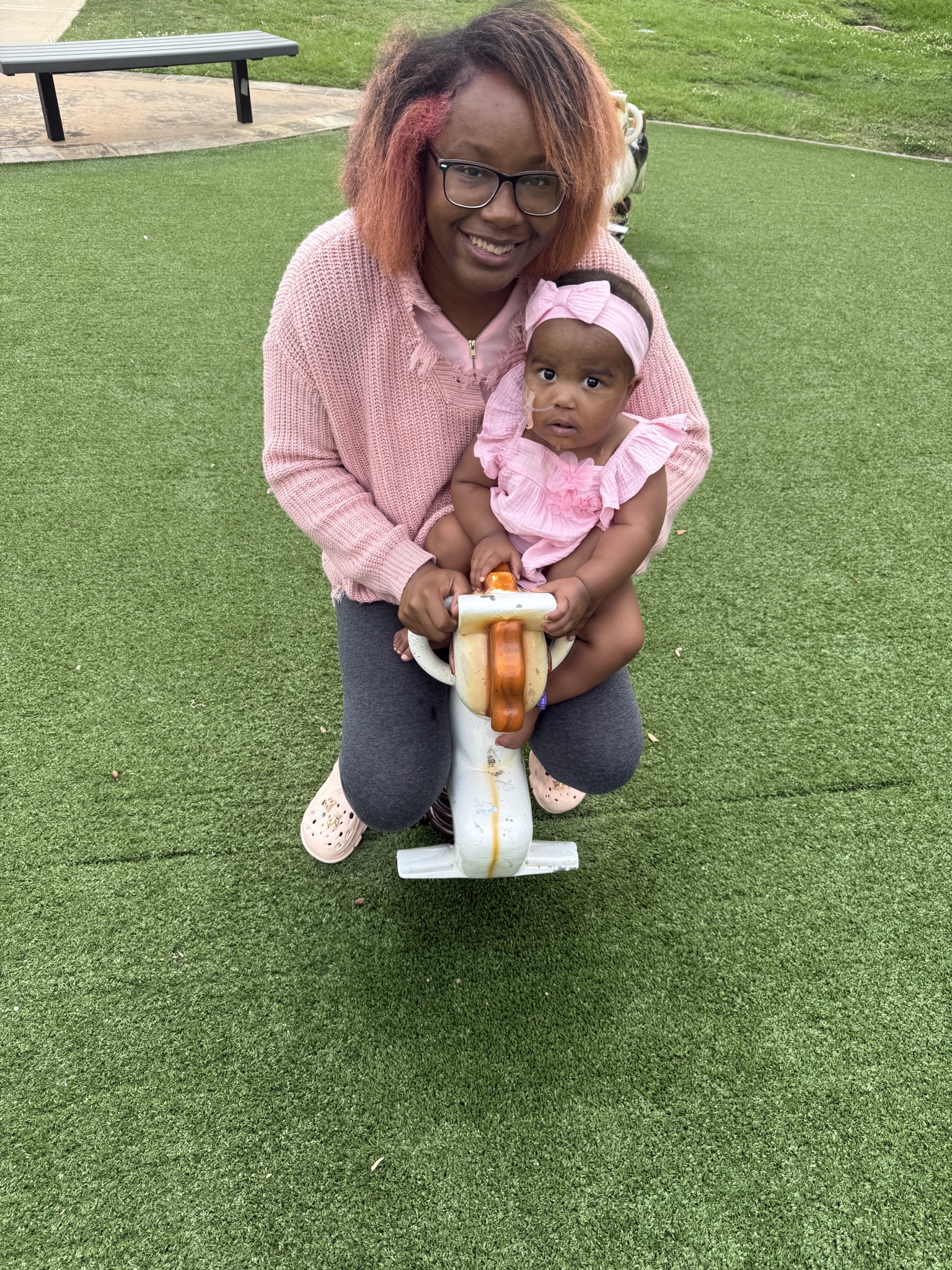 Toddler Kylin smiling on a playground spring rider with her mother Diamond sitting behind her on green turf.