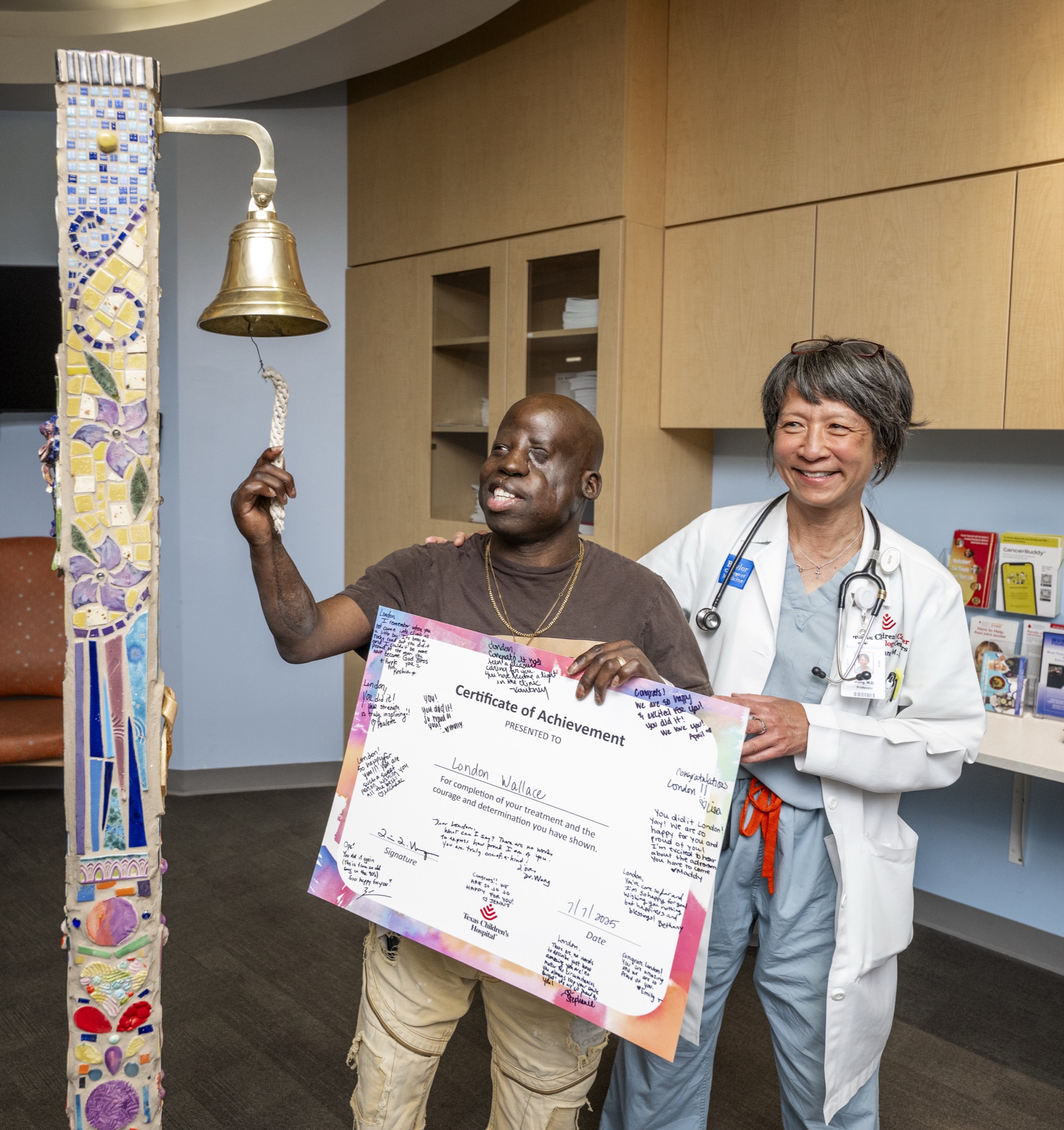 London Wallace ringing the ceremonial treatment completion bell while holding a signed Certificate of Achievement next to his oncologist Dr. Lisa L. Wang at Texas Children's Hospital.